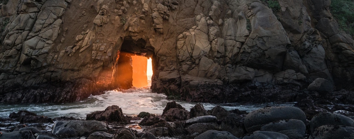 Klippa vid hav som har en tunnel öppning formad som en rektangel där solen lyser igenom